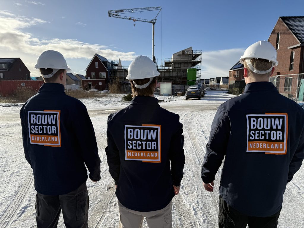 Drie vakmensen in werkjassen met logo van Bouwsector Nederland kijken naar rijen woningen in aanbouw op een winterdag